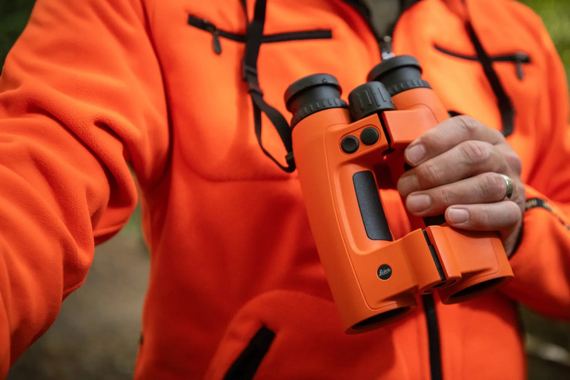 Person in orange jacket holding a pair of orange binoculars in a forest setting.